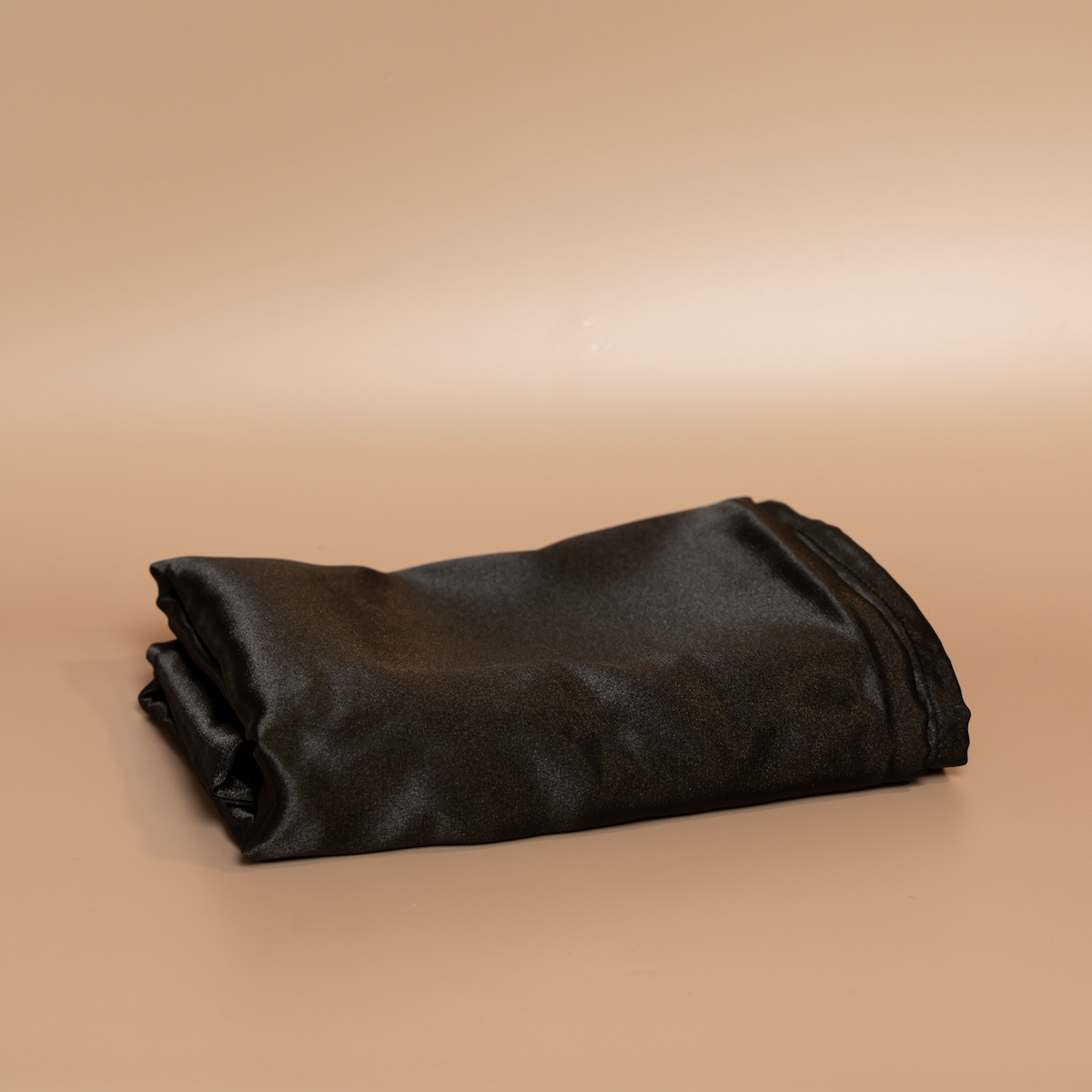 Folded black ROYAL silk satin pillowcase photographed against a beige background.