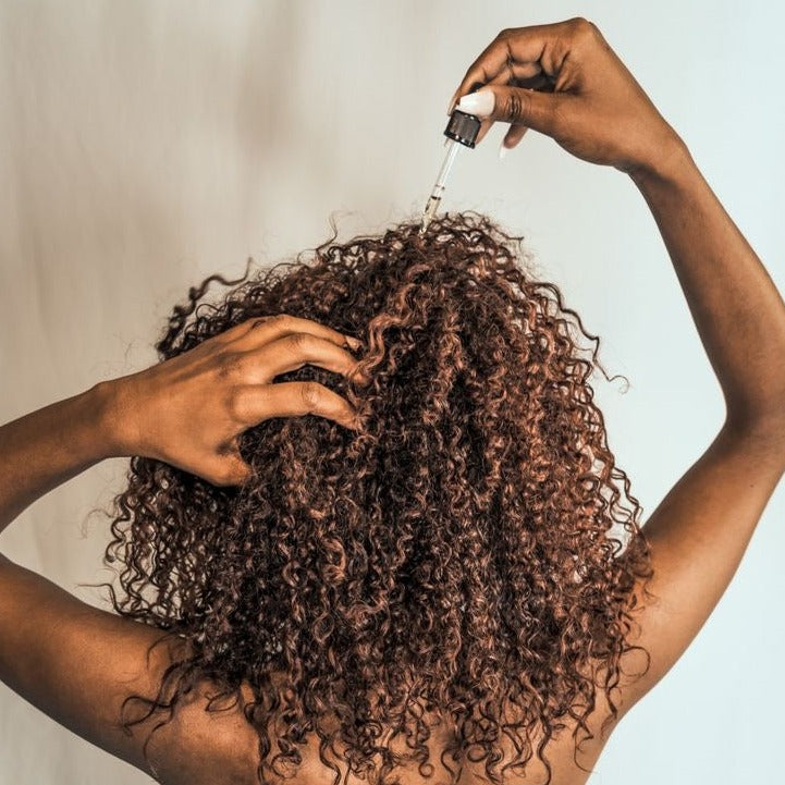 Curly hair shown during targeted application of a dropper serum directly onto the scalp.