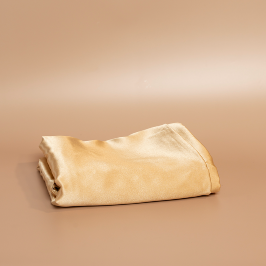 Folded gold ROYAL silk satin pillowcase photographed against a beige background.