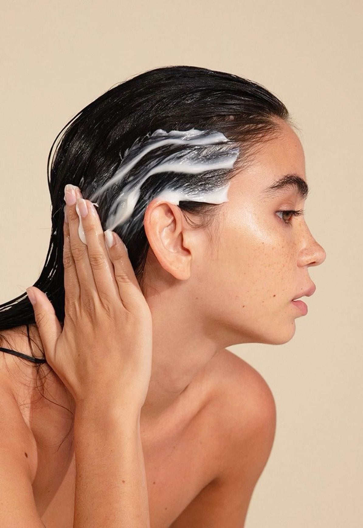 Woman applying a white hair care product to her dark hair, shown in profile against a beige background.