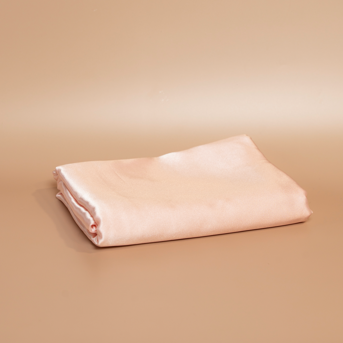 Folded light pink ROYAL silk satin pillowcase photographed against a beige background.