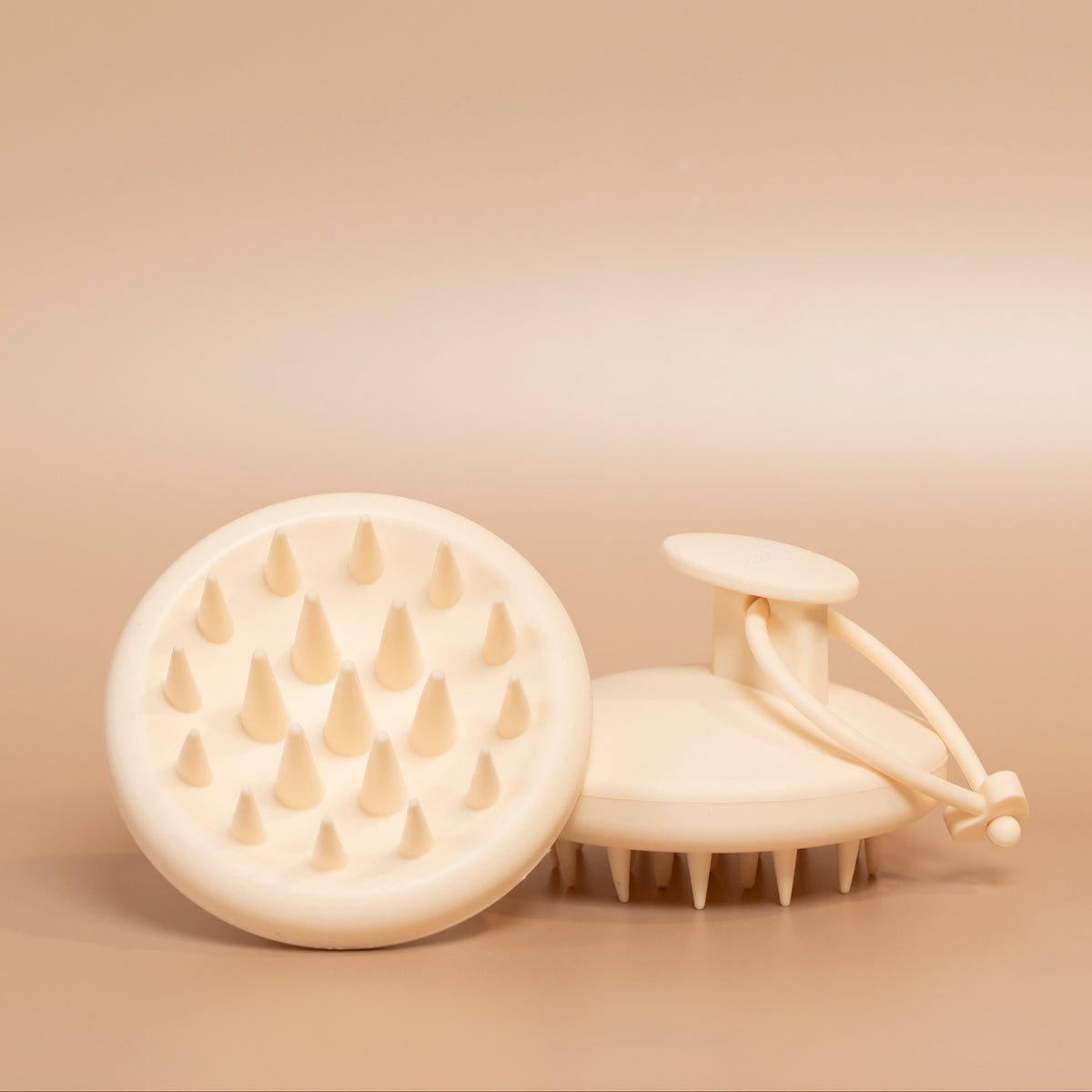 Two cream-colored silicone scalp massage brushes, one showing the flexible bristles and the other showing the handle, on a warm beige background.
