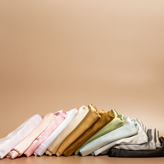 ROYAL silk satin pillowcases in a variety of colours, folded and arranged together against a beige background.