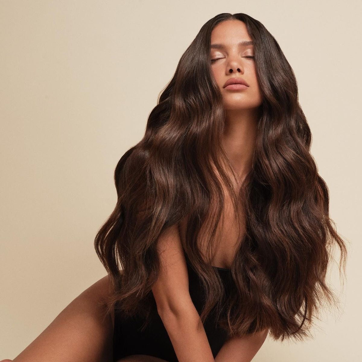 Woman with long, voluminous brown hair styled in soft waves against a neutral beige background.